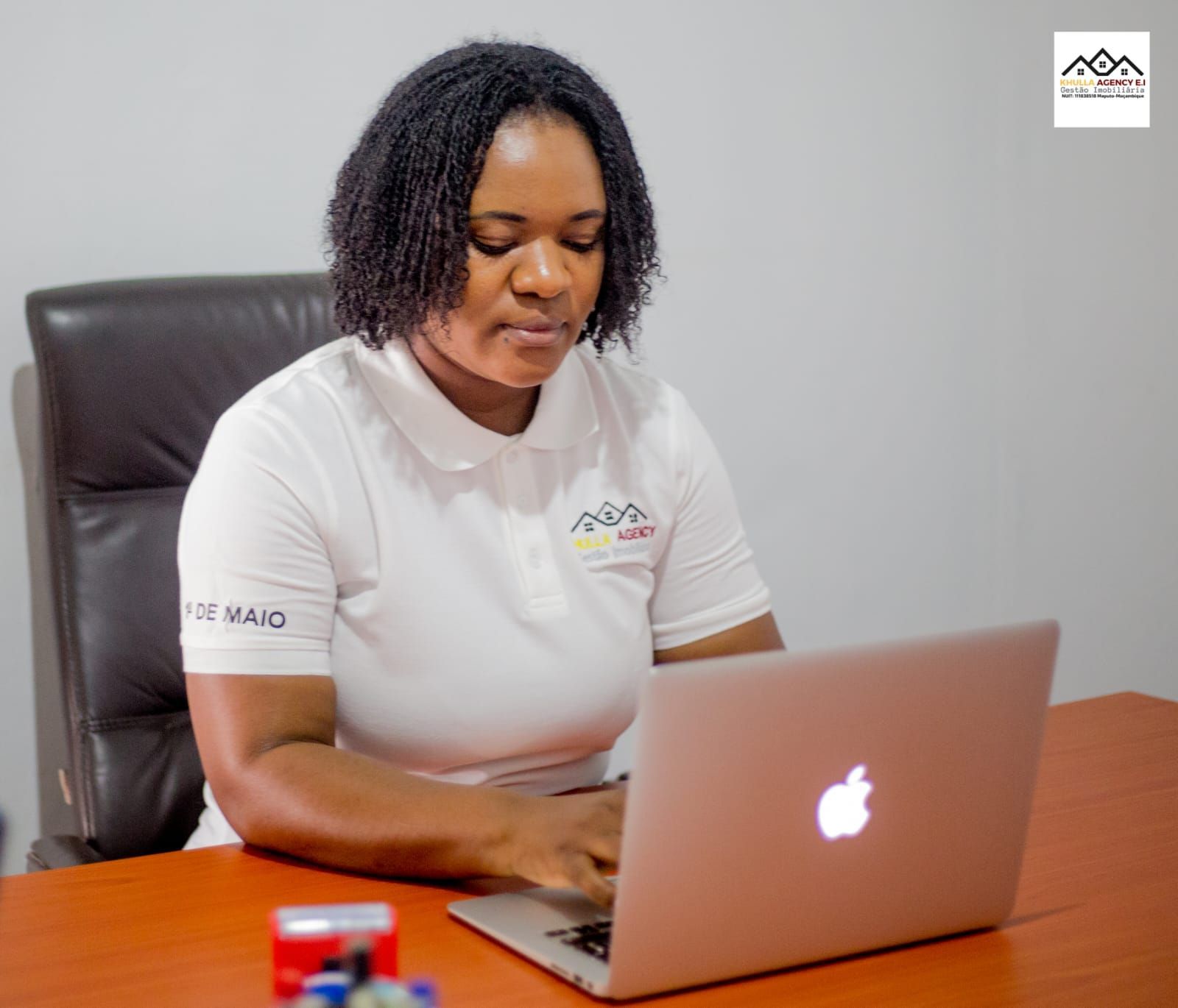 Woman typing on a laptop at a desk in an office setting. She wears a white polo shirt.