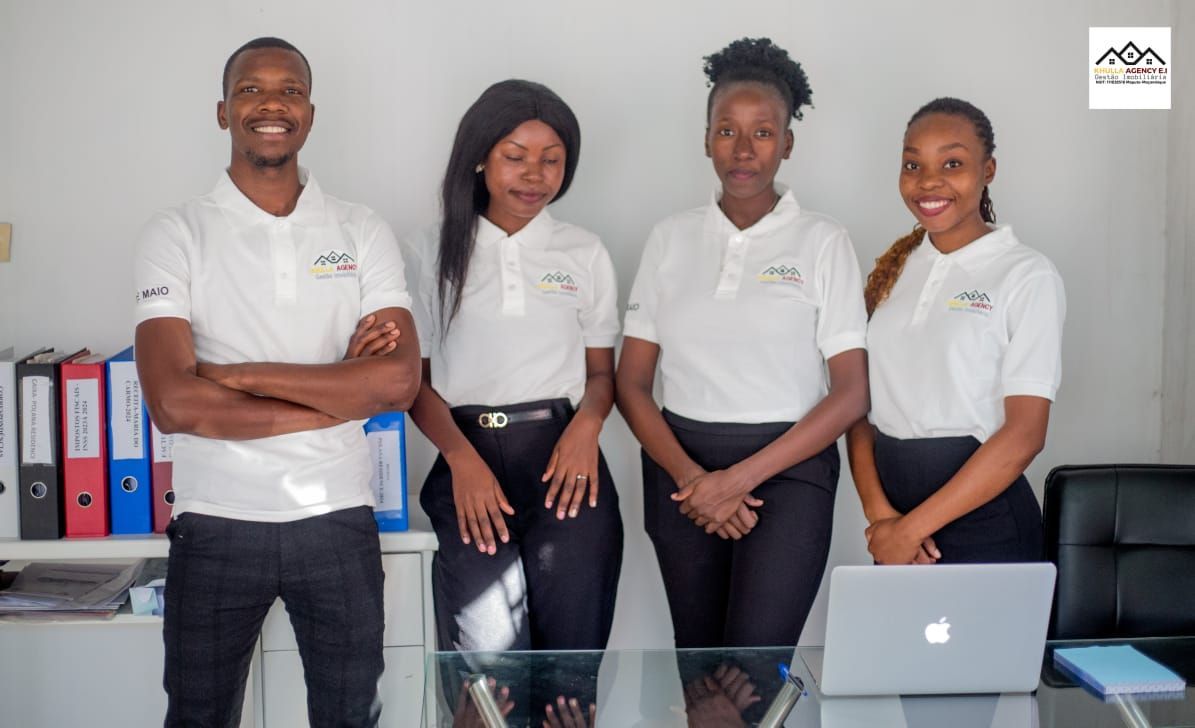 Four people in white shirts and black pants standing behind a desk with a laptop.
