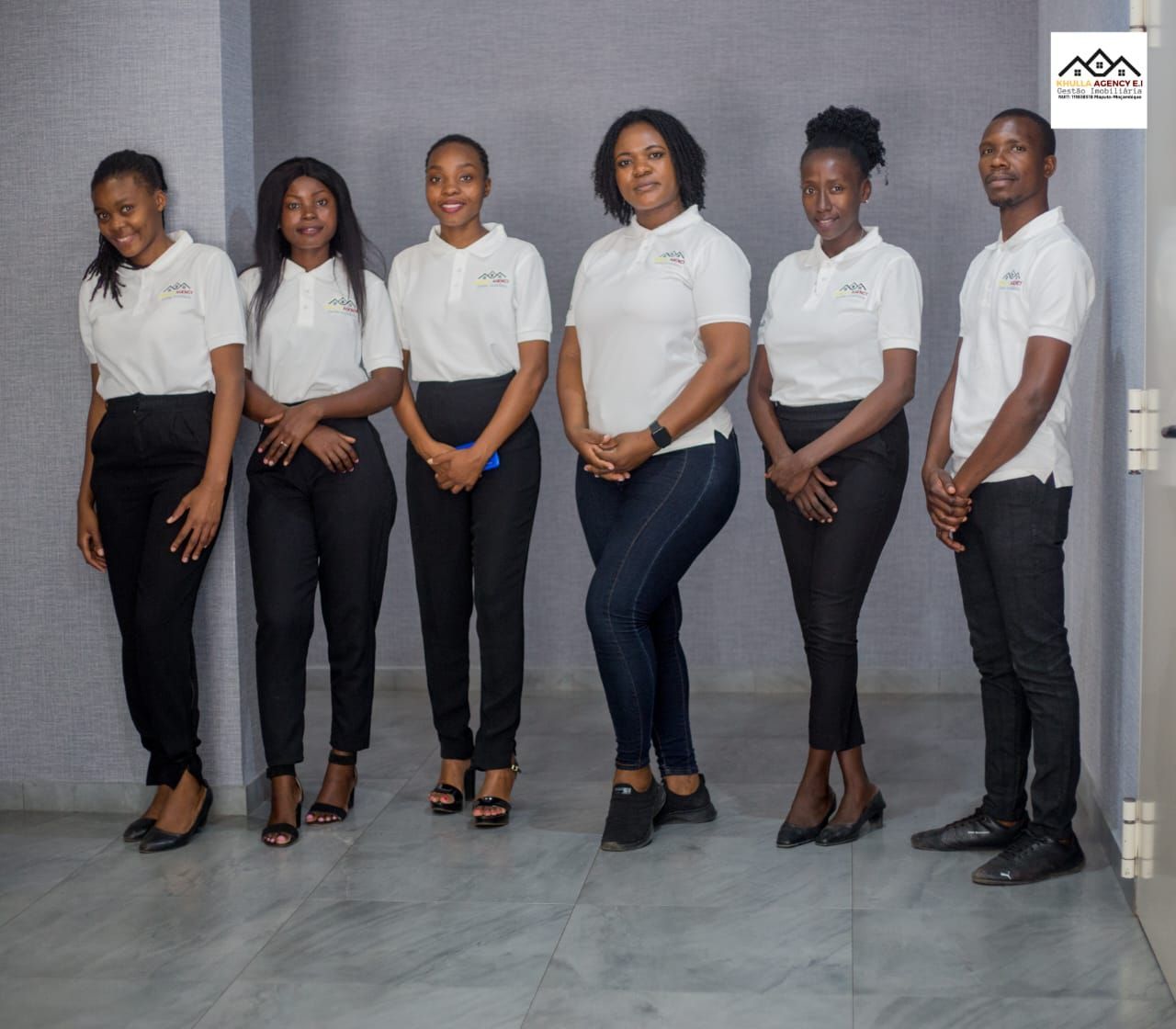 Six people, in white shirts and dark pants, stand in a room. Some are smiling.
