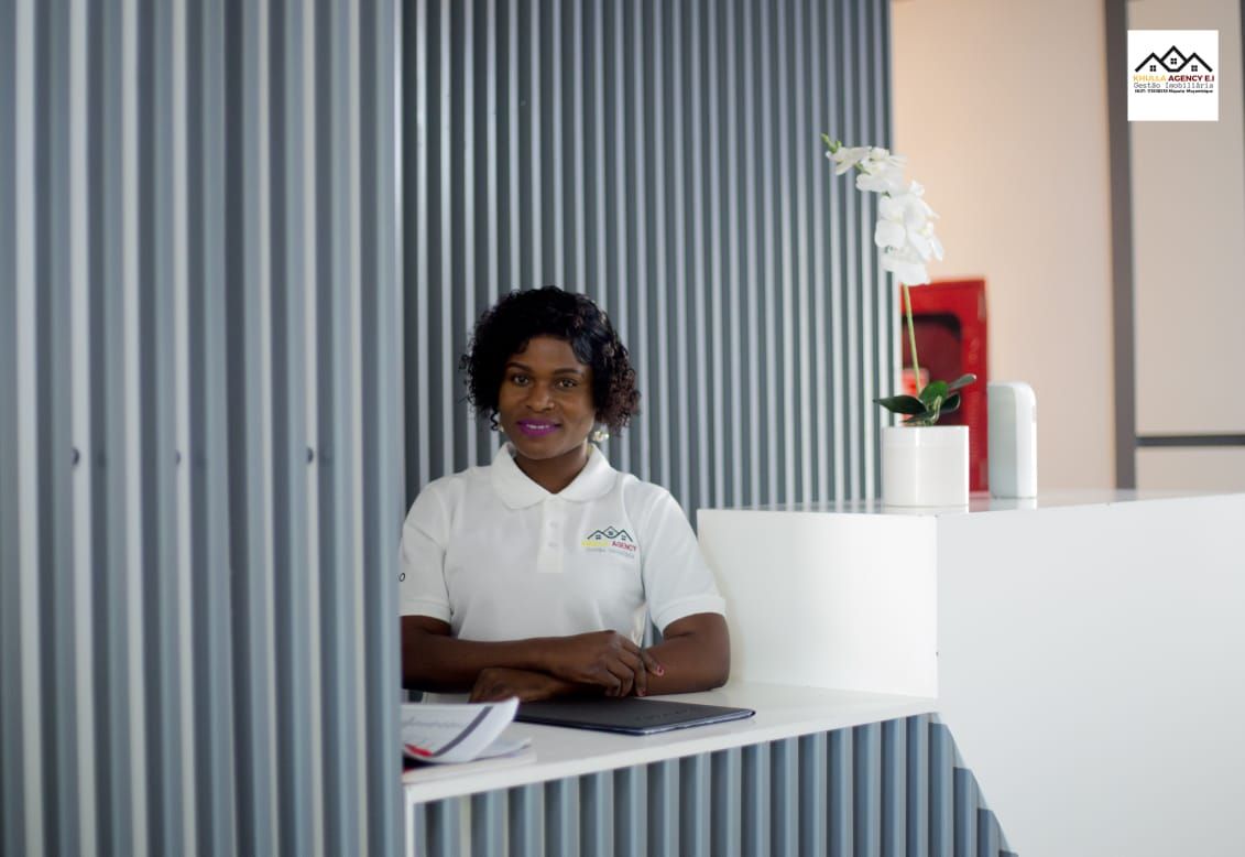 Woman at a modern white reception desk, smiling.  Gray corrugated wall. White shirt. Plant and decorations.