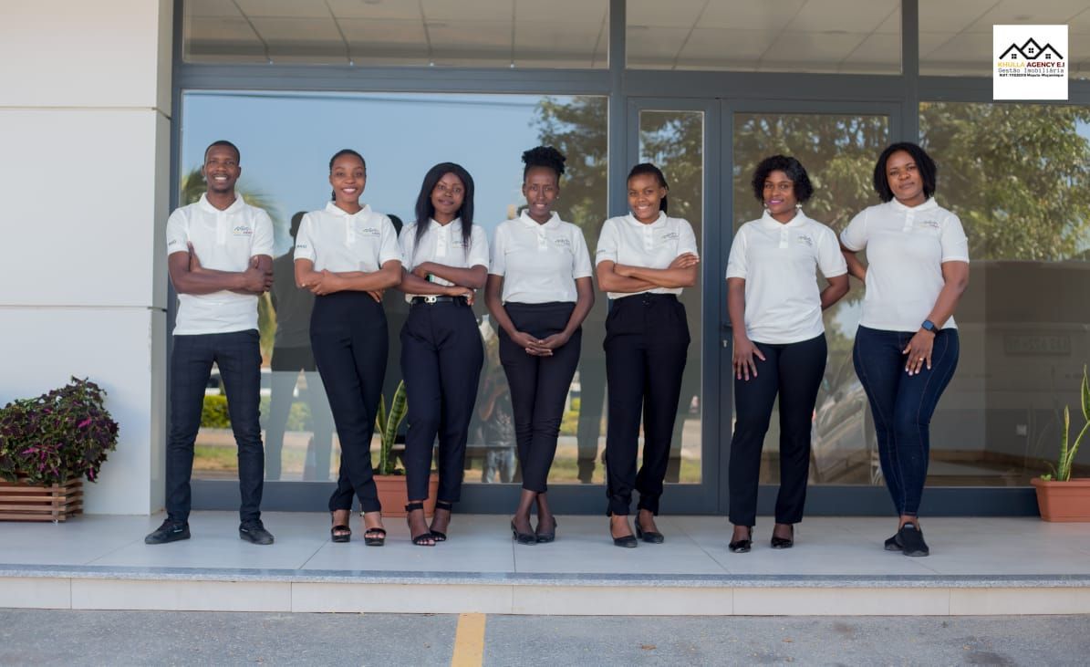 Group of seven people in matching white shirts and black pants, standing in front of a building.
