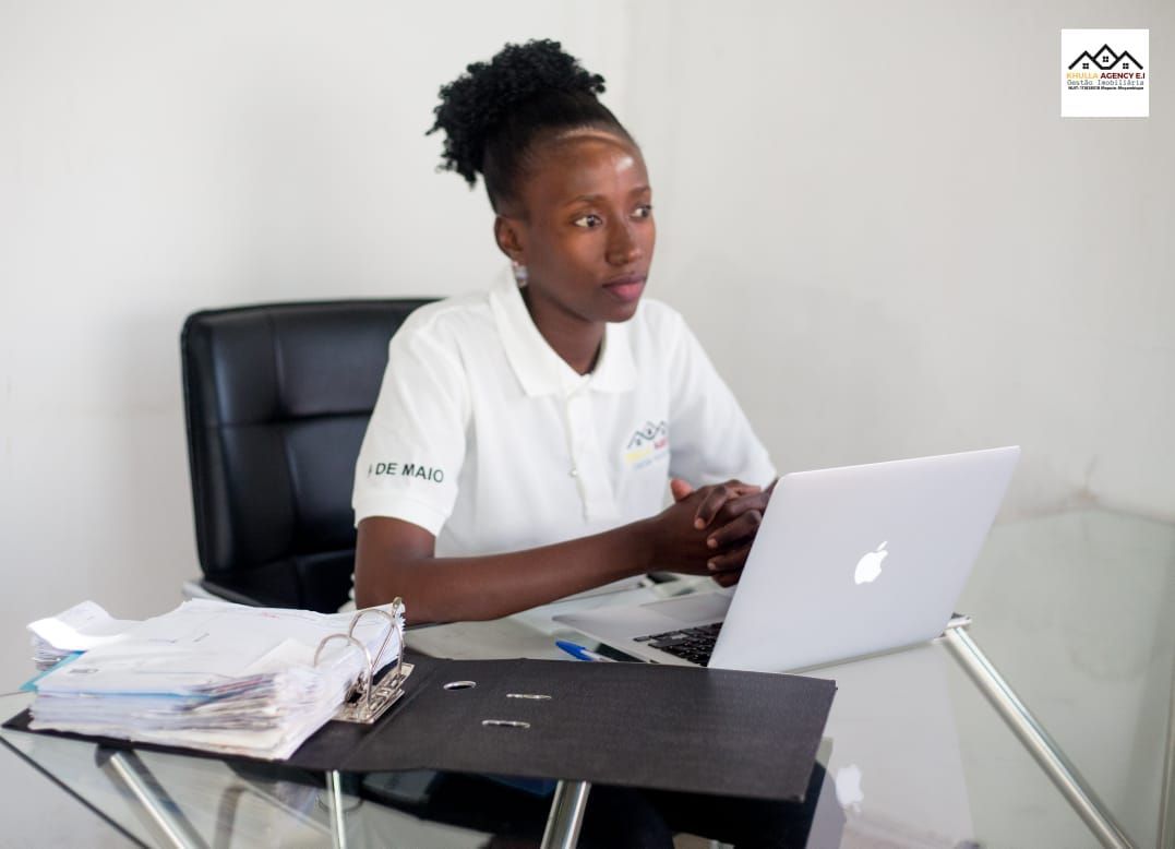 Woman at a desk with laptop, papers, and pen. She wears a white shirt and has a focused expression.