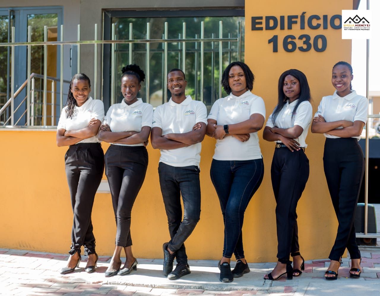 Group of six people in front of a building, arms crossed. White shirts, dark pants. Building is yellow, says