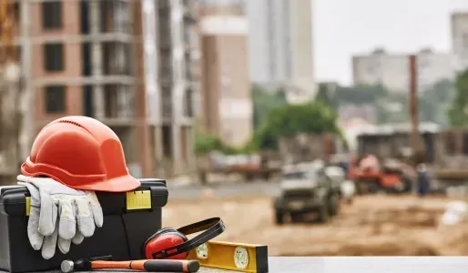 Orange hard hat, gloves, and tools on a toolbox at a construction site, with buildings in the background.