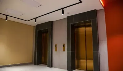 Two elevators with gold doors in a building's hallway; tan and orange walls, black track lighting.