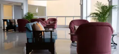 A lounge area with burgundy armchairs and a blue couch. Large windows, shiny floors, and potted plants.