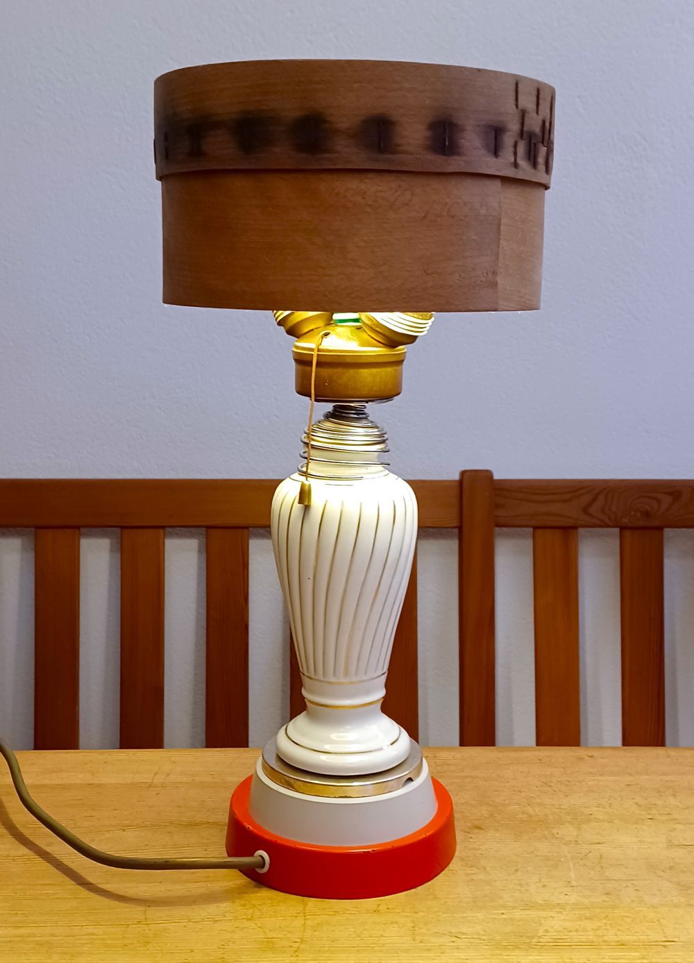Tischlampe mit weißem Fuß, rotem Ständer und braunem Trommelschirm auf einem Holztisch