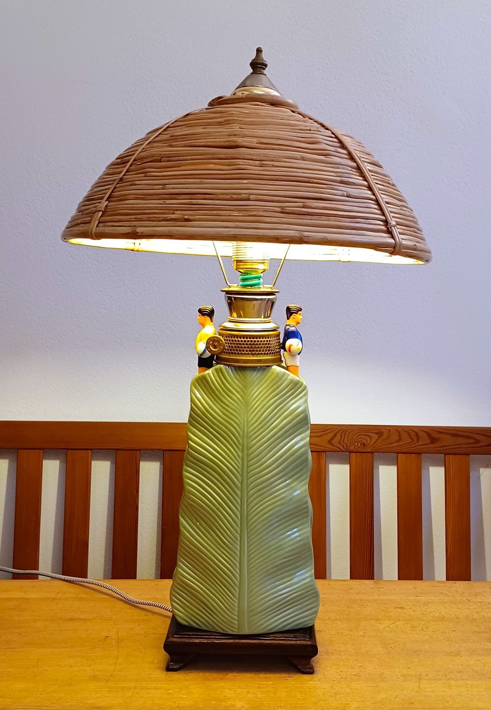 Gewebte Tischlampe mit breitem Strohschirm und grünem, strukturiertem Fuß auf einem Holztisch