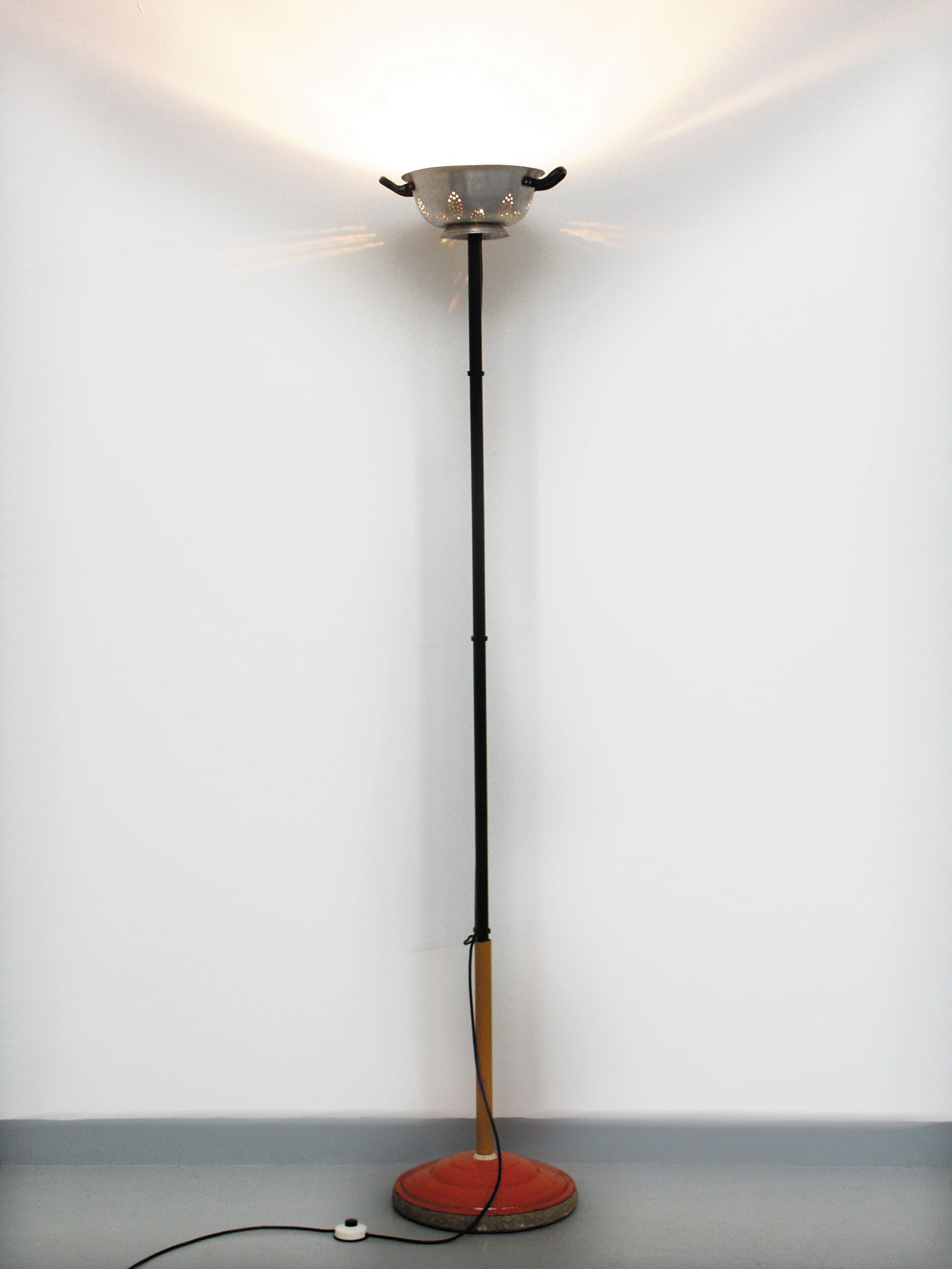 Stehlampe mit rotem Fuß und schwarzem Ständer vor einer weißen Wand in einem spärlich möblierten Zimmer