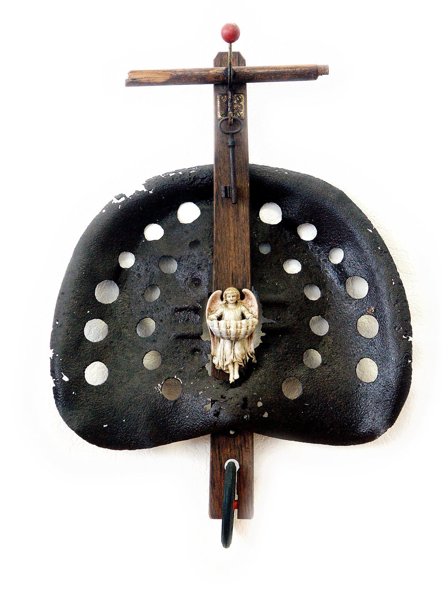 Dekoratives Holzkreuz mit einem kleinen Totenkopf, montiert auf einer schwarzen, muschelförmigen Plakette mit weißen Punkten.