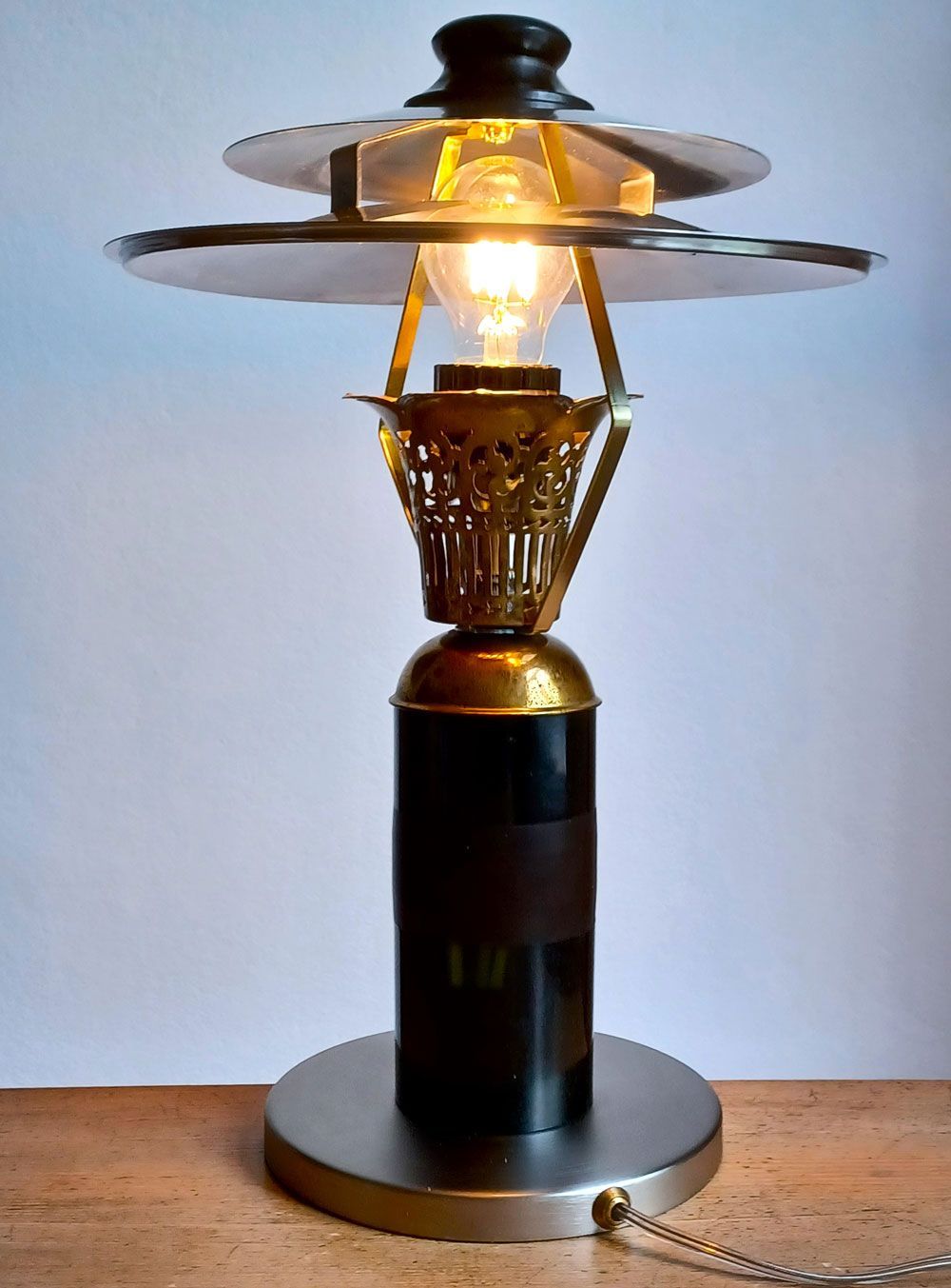 Tischleuchte mit leuchtender Glühbirne, schwarzem Sockel und Metallschirm auf einer Holzoberfläche