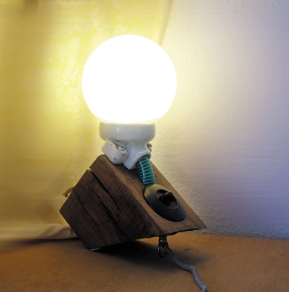 Eine leuchtende Glühbirne ist mit einer Kette und einem Kabel an einem Holzblock befestigt und hängt an einer schlichten Wand.