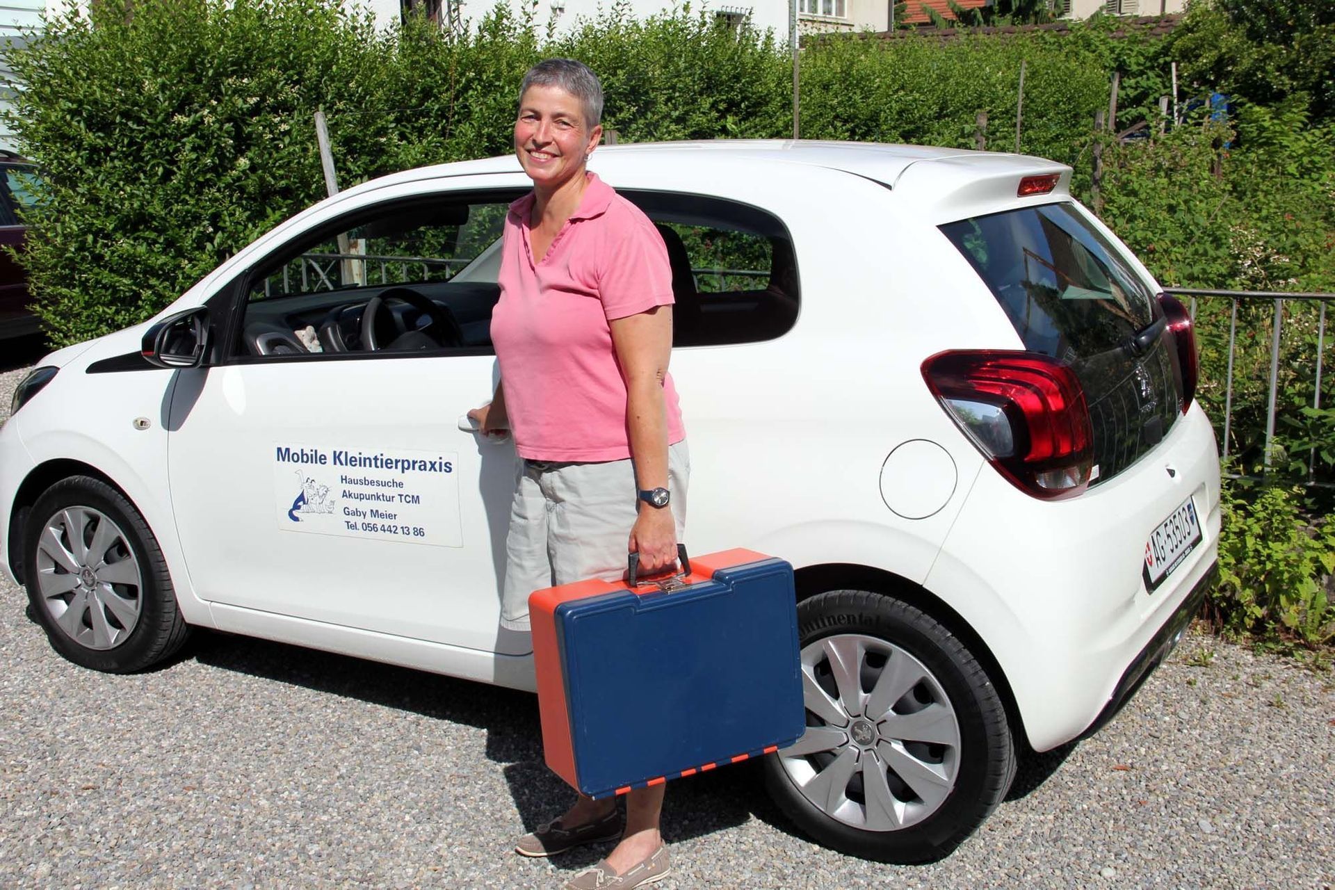 Frau steht neben einem weissen Auto und hält einen blau-orangenen Koffer; im Freien.