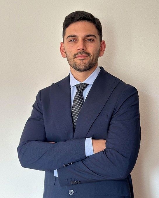 Hombre con traje azul, con los brazos cruzados y sonriendo ligeramente, frente a una pared blanca.