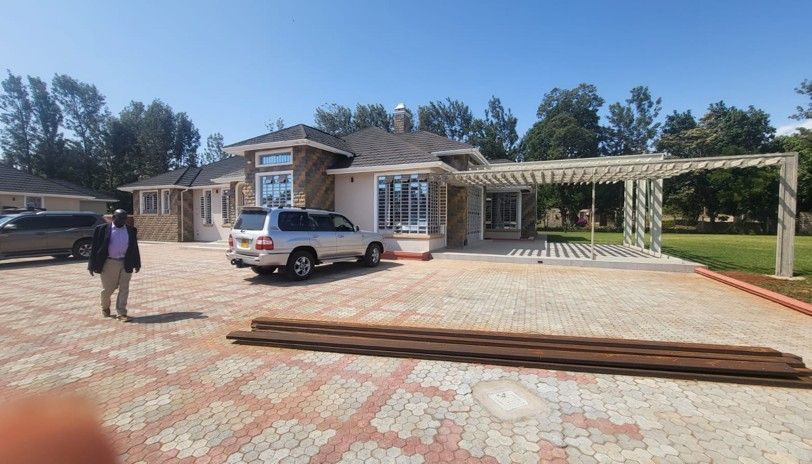 A person stands in front of a house with a car parked in the driveway. A pergola is attached to the house.