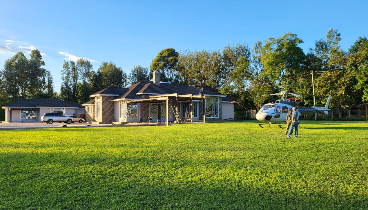 Helicopter lands on a green lawn in front of a house. Person walks toward the helicopter.
