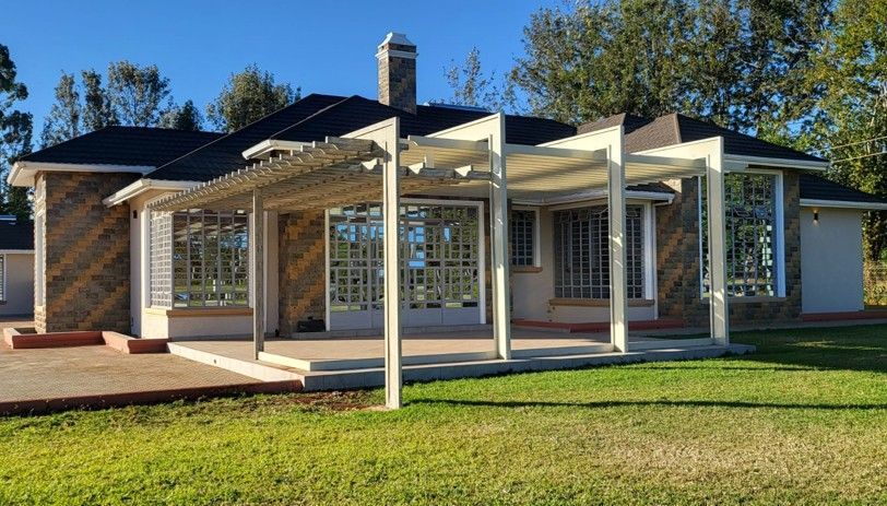 House with a pergola and stone exterior, surrounded by green grass and trees under a clear blue sky.