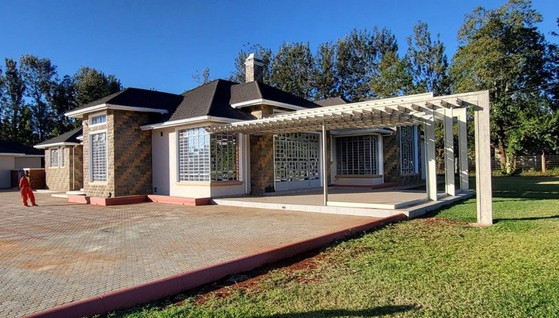 House with stone facade and pergola, on a paved driveway and lawn, under a blue sky.