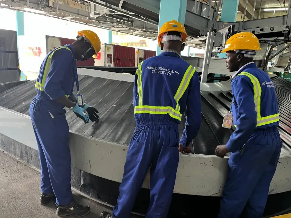 Three men wearing hard hats and safety vests are working on a conveyor belt.