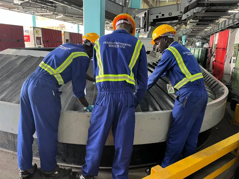 Three men in blue overalls and yellow hard hats are working on a conveyor belt