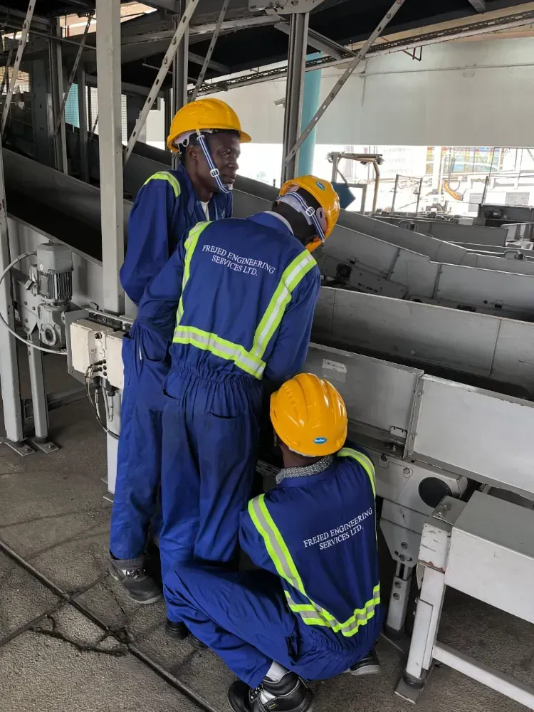 Three men wearing hard hats and blue overalls are working on a conveyor belt.
