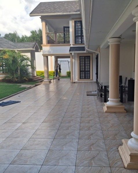 A large, light-colored house with a tiled patio and several columns. Green grass and trees are in the background.