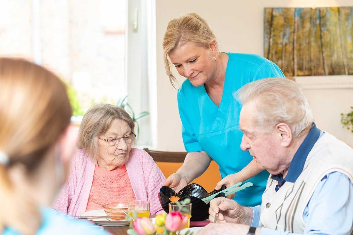 Eine Krankenschwester serviert einer Gruppe älterer Menschen, die an einem Tisch sitzen, Essen.