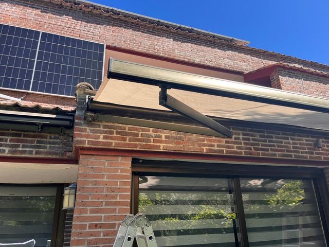 Fachada de ladrillo con paneles solares en el tejado y un toldo retráctil beige sobre un patio.