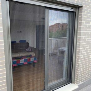 Puerta corrediza de cristal con mosquitera negra, que deja ver el salón y el balcón.