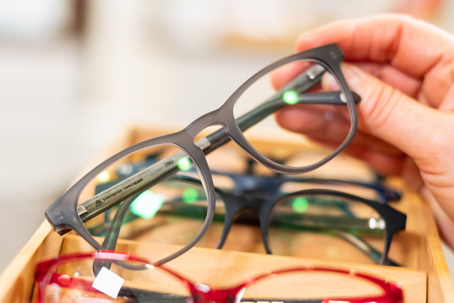 Eine Hand hält eine schwarze Brille über einer Schachtel mit verschiedenen Brillengestellen.