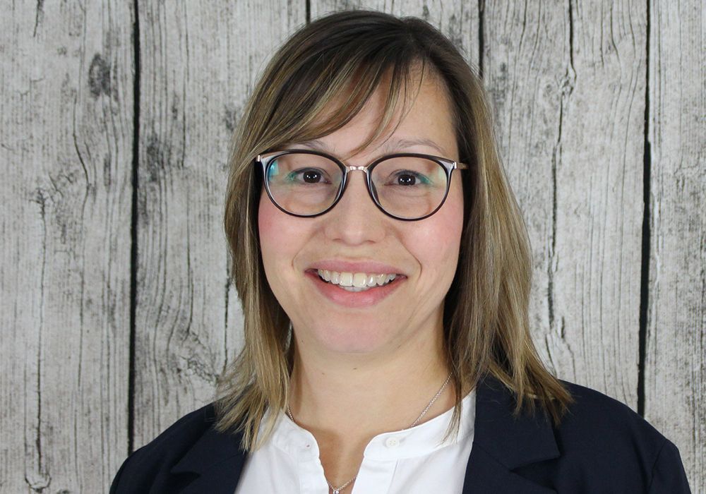 Lächelnde Person mit Brille und dunklem Blazer vor einer grauen Holzwand