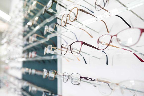 Reihen von Brillen, die in einem Optikergeschäft an einer Wand ausgestellt sind, mit verschiedenen Gestellen in Silber, Schwarz und Rot.