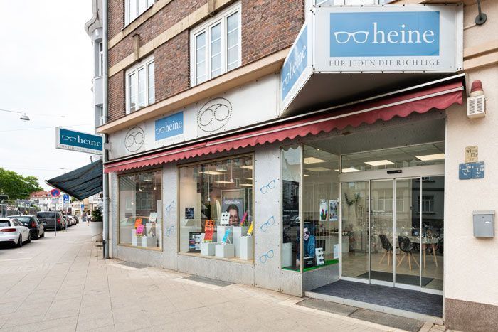 Optikergeschäft mit blauem Schild, roter Markise und gläsernem Eingang an einer Stadtstraße