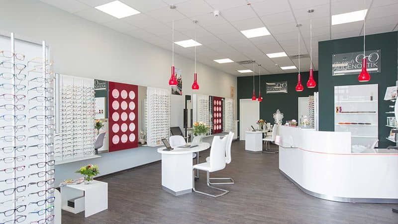 Heller, moderner Brillen-Showroom mit weißen Auslagen, roten Pendelleuchten und Beratungsstühlen