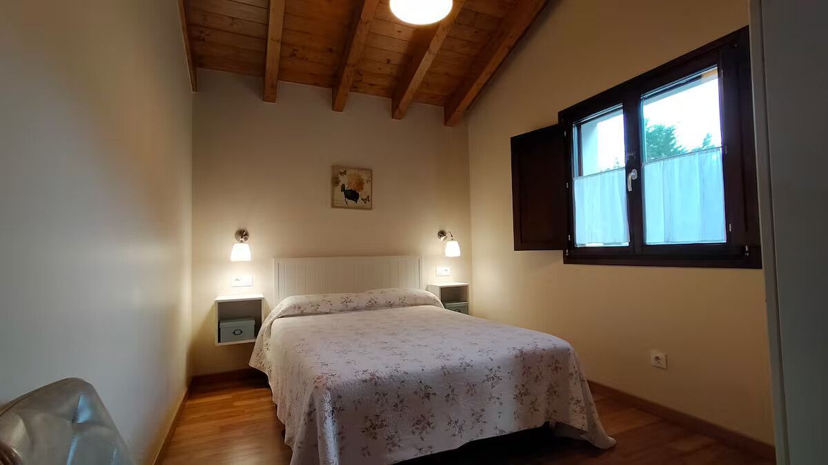 Acogedor dormitorio con una cama, techo con vigas de madera, lámparas de mesita de noche y una ventana con marco oscuro