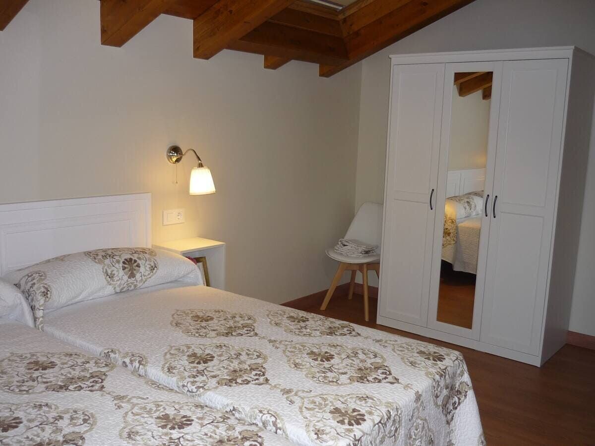 Dormitorio con cama blanca, armario con espejo, silla y vigas de madera en el techo.