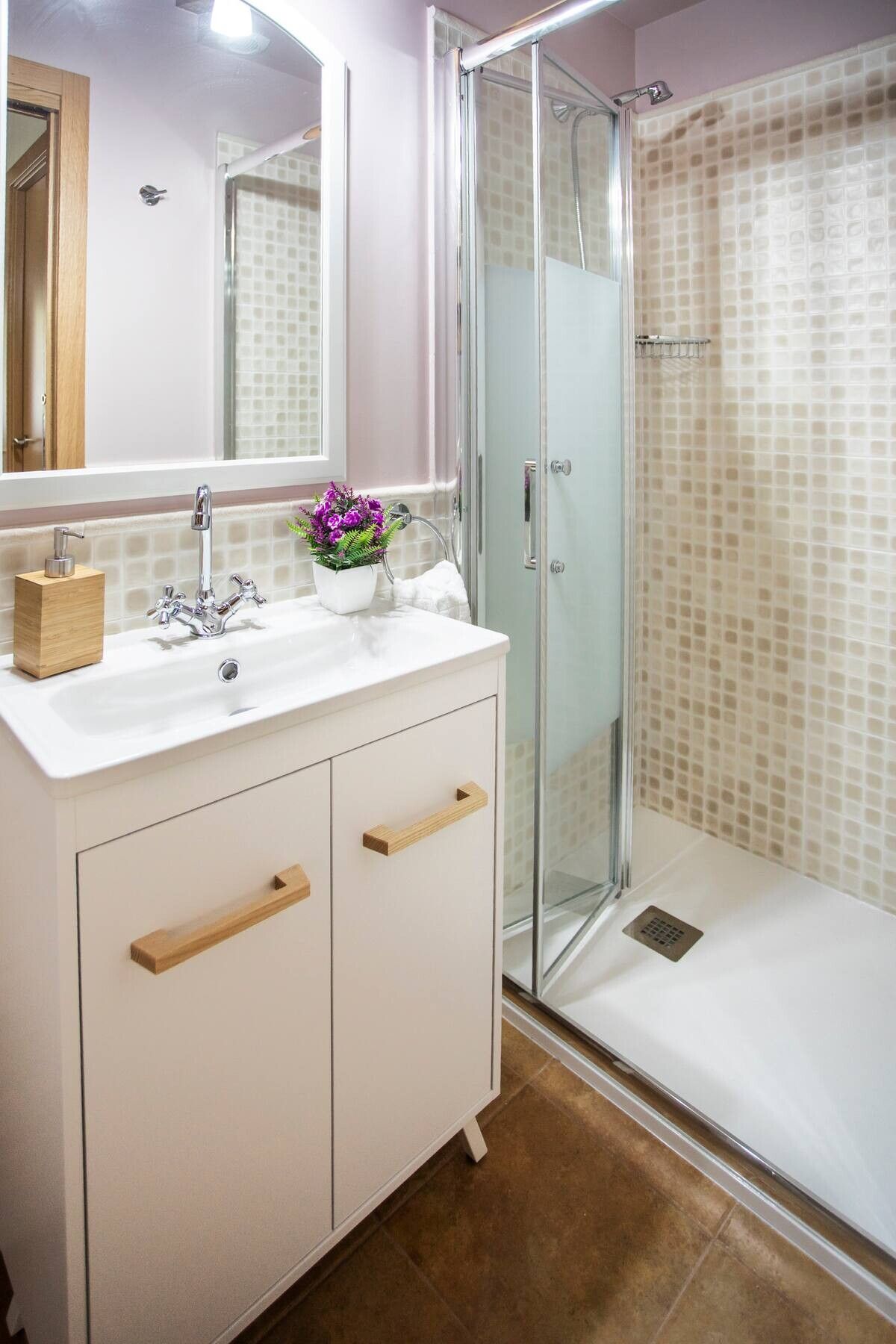 Baño compacto con mueble de lavabo blanco, espejo y ducha con mampara de vidrio y paredes revestidas con azulejos.