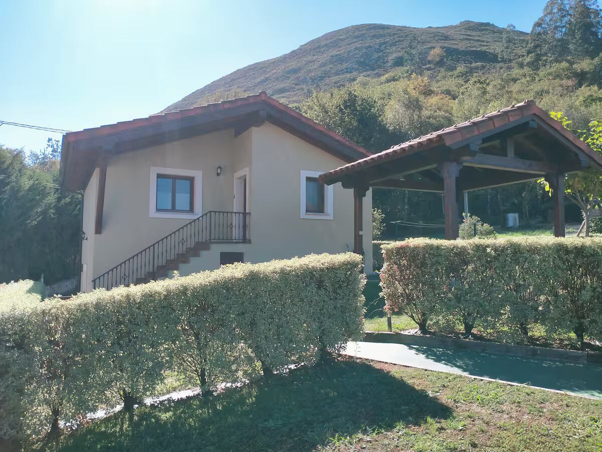Casa pequeña con tejado rojo junto a un seto y un patio cubierto, con una ladera verde al fondo bajo un cielo azul