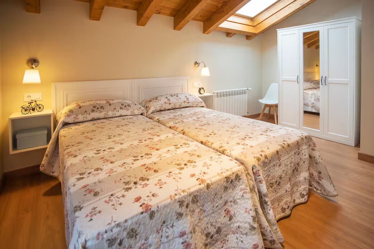 Acogedor dormitorio abuhardillado con colcha floral, vigas de madera a la vista y puertas del armario abiertas