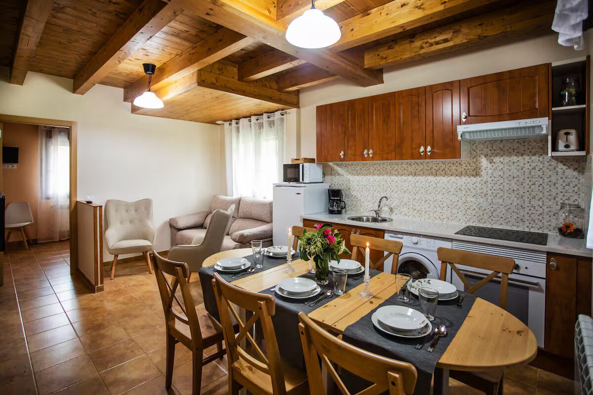Acogedor comedor y cocina de estilo rústico, con vigas de madera, una mesa redonda preparada para cuatro personas y un salpicadero de azulejos.