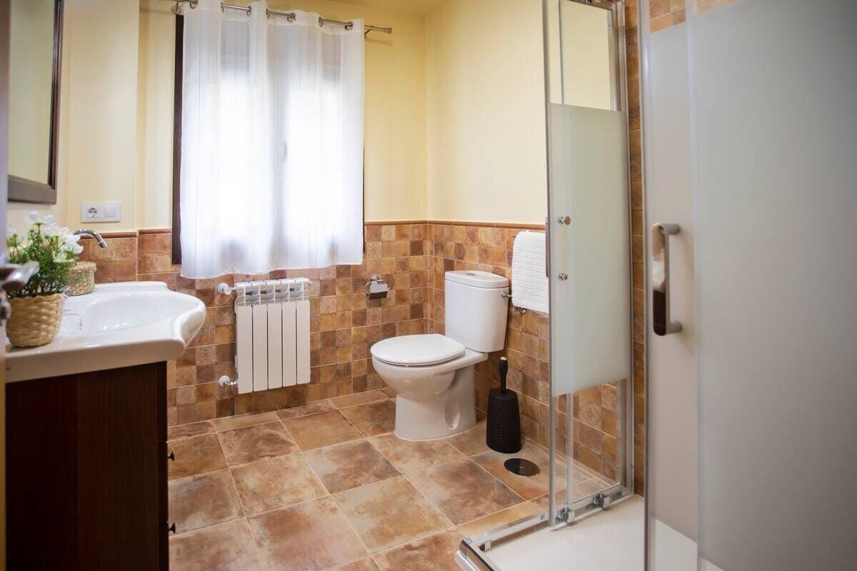 Baño luminoso con lavabo, inodoro, ducha y ventana con cortinas blancas