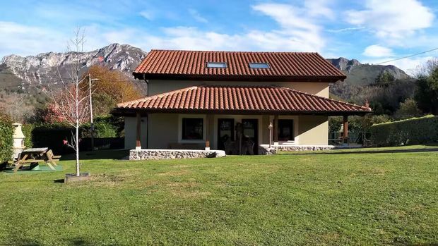 Casa de dos plantas con tejado de tejas rojas y césped verde, rodeada de montañas bajo un cielo azul.