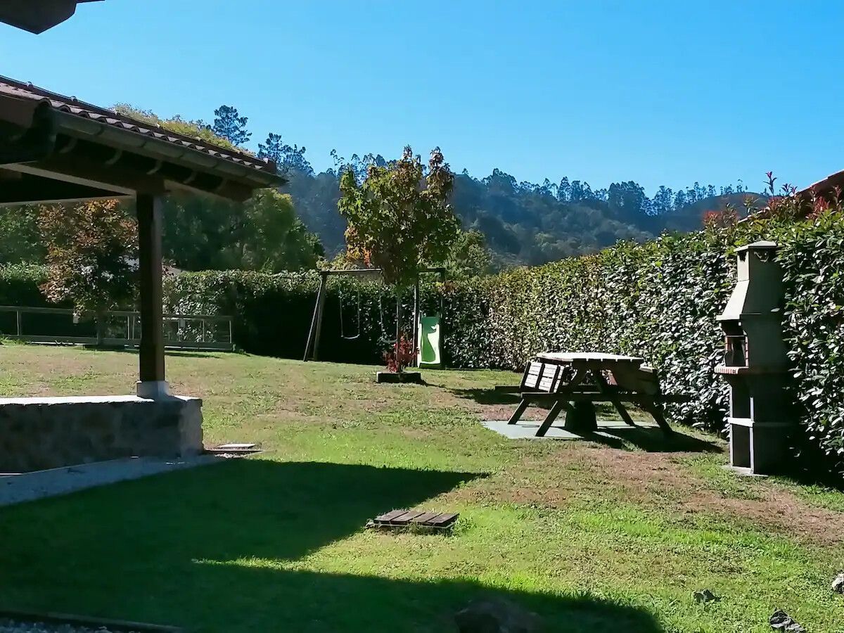 Patio trasero soleado con una mesa de pícnic, un muro de piedra, césped y árboles bajo un cielo despejado