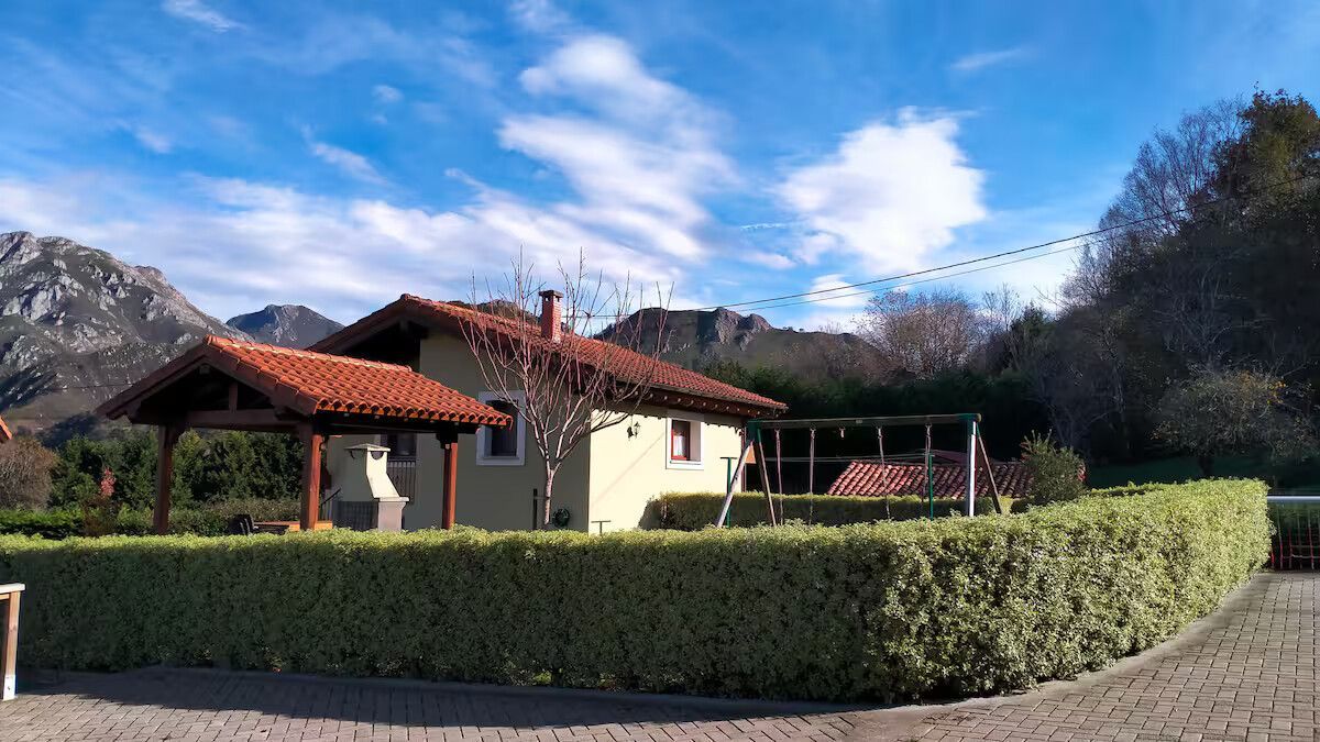 Casa de campo con tejado de tejas rojas, seto recortado y montañas bajo un cielo azul
