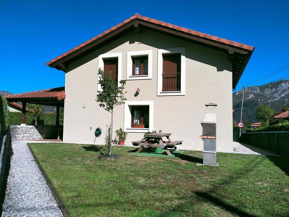 Casa de dos pisos de color crema, con techo rojo, césped verde, mesa de pícnic y montañas al fondo