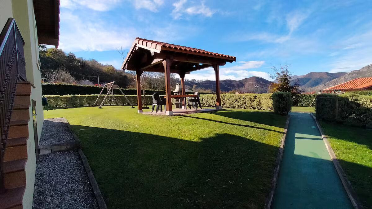 Patio soleado con césped verde, pérgola de madera y vistas a la montaña junto a un sendero bordeado de setos