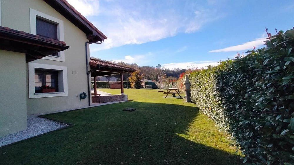 Casa con un césped verde a la sombra, un seto como cerca y montañas a lo lejos bajo un cielo azul