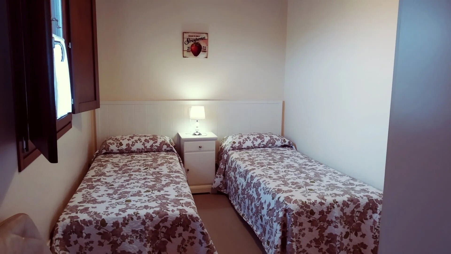 Dormitorio pequeño con dos camas individuales, colchas de flores y una mesita de noche con una lámpara entre ellas.