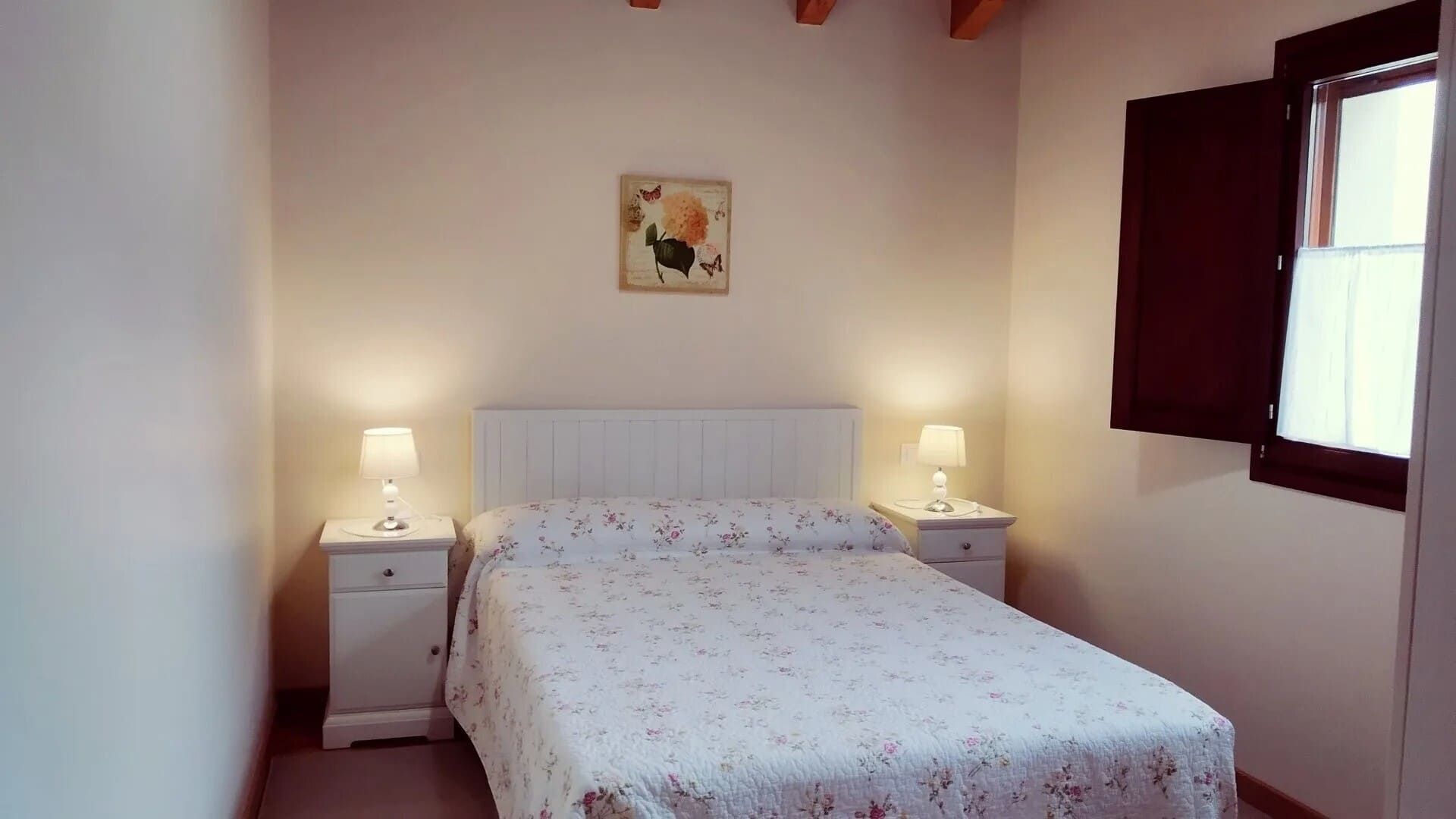 Dormitorio pequeño con una cama, dos lámparas de mesilla, un cuadro en la pared y una ventana con cortinas oscuras