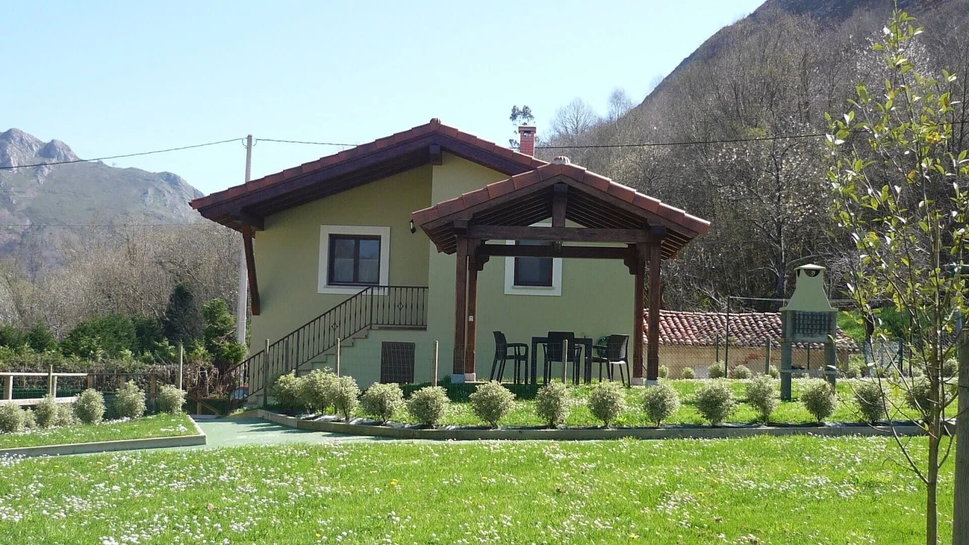 Casita de color crema con tejado rojo en un jardín con césped, rodeada de árboles y colinas.